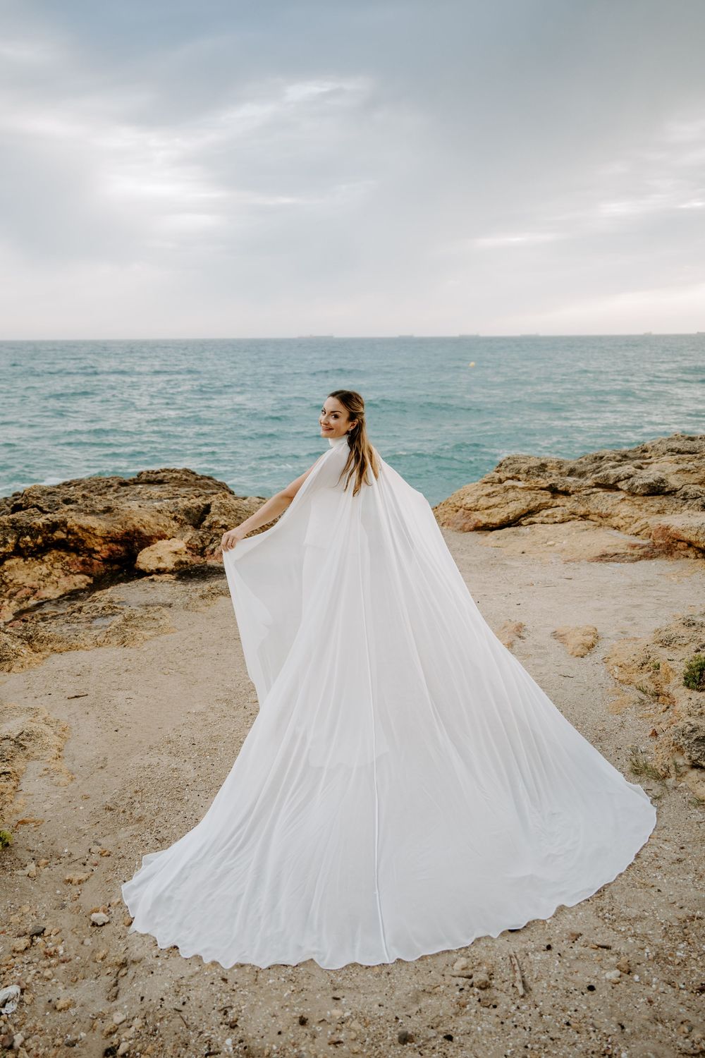Bride in a flowing white gown and long cape looks over her shoulder on a rocky seaside cliff overlooking the turquoise sea.
