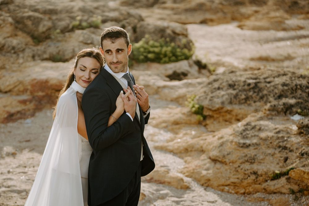 Bride in a white veil and groom in a dark suit embrace on a rocky coastal shoreline during a romantic outdoor wedding photoshoot.