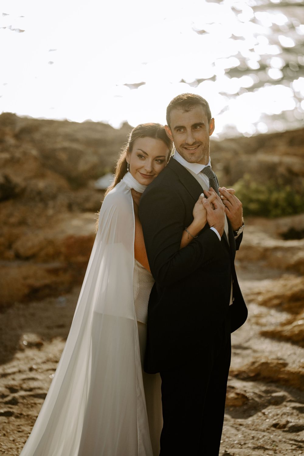 Smiling bride in a flowing white cape embraces her groom in a dark suit from behind on sunlit coastal rocks.