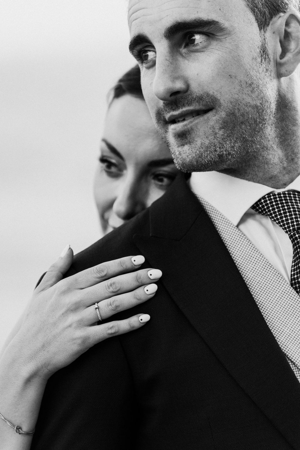 Black and white portrait of a smiling bride in a ring and white manicure embracing a groom in a suit from behind.