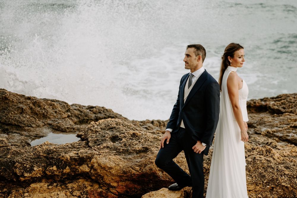 Bride in a flowing white gown and groom in a dark suit pose on rocky coastal cliffs as ocean waves crash behind them.