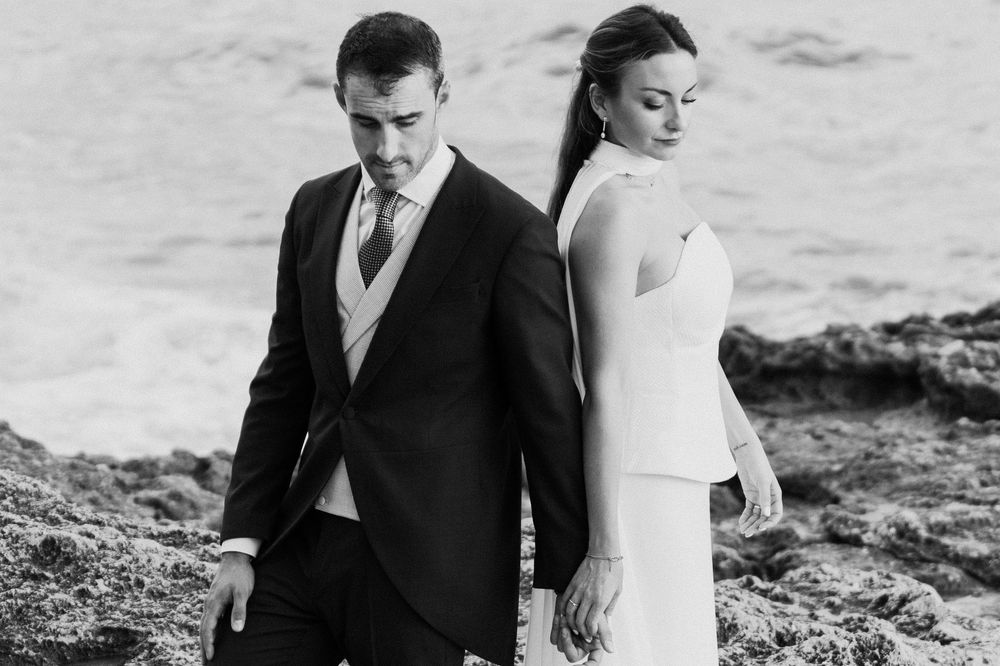 Black and white photo of a groom in a suit and a bride in a sleek white dress standing back to back on rocky shoreline by the sea.