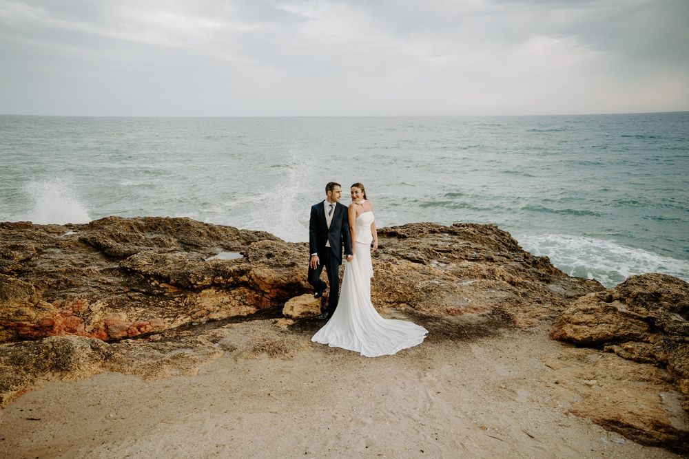 Bride in a white gown and groom in a dark suit pose together on rocky coastal cliffs with ocean waves behind them.