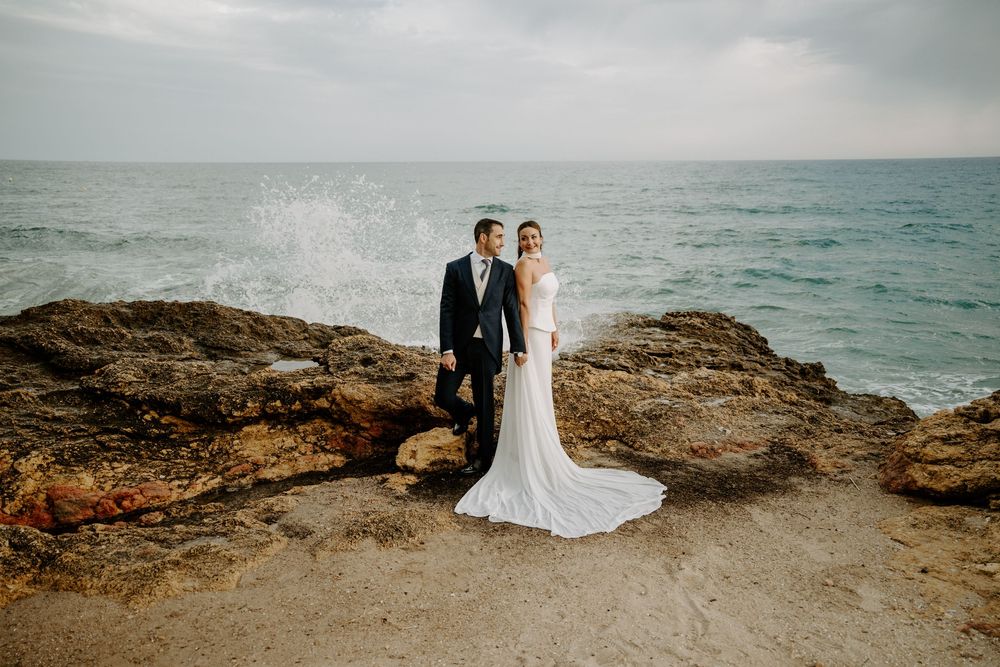 Bride in a long white gown and groom in a dark suit stand on rugged seaside rocks as ocean waves splash behind them.