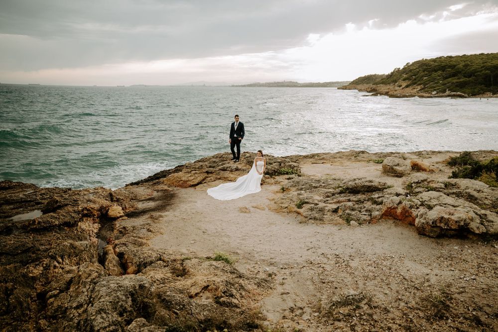 Bride in a flowing white gown and groom in a dark suit stand on a rocky seaside cliff overlooking the sea at sunset.