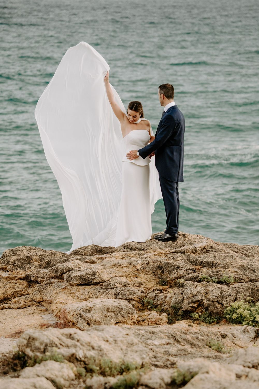 Bride in a flowing white gown and veil stands with her groom in a navy suit on rocky seaside cliffs overlooking the turquoise ocean.