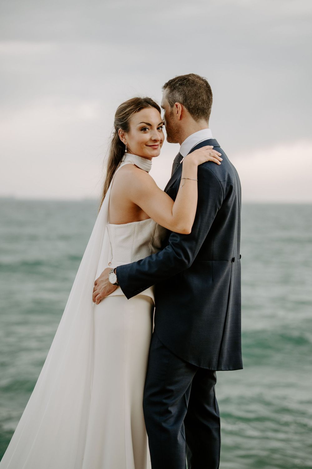 Bride in a white gown with a long veil smiles over her shoulder while embracing her groom in a navy suit by the sea.