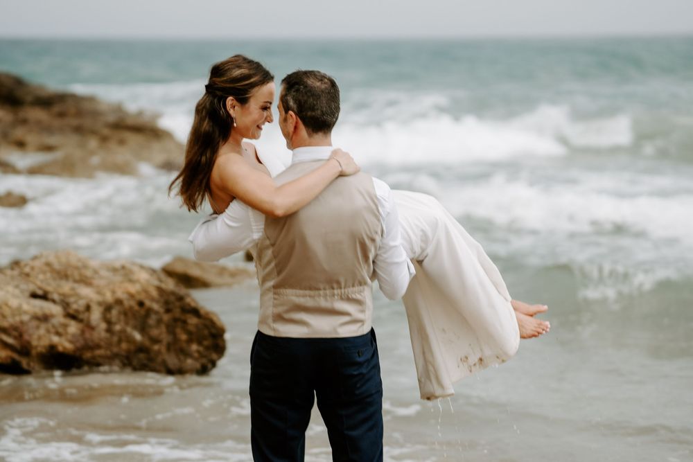 Groom in a vest carries his bride in a white dress through shallow ocean waves beside rocky shoreline during a beach wedding photo shoot.