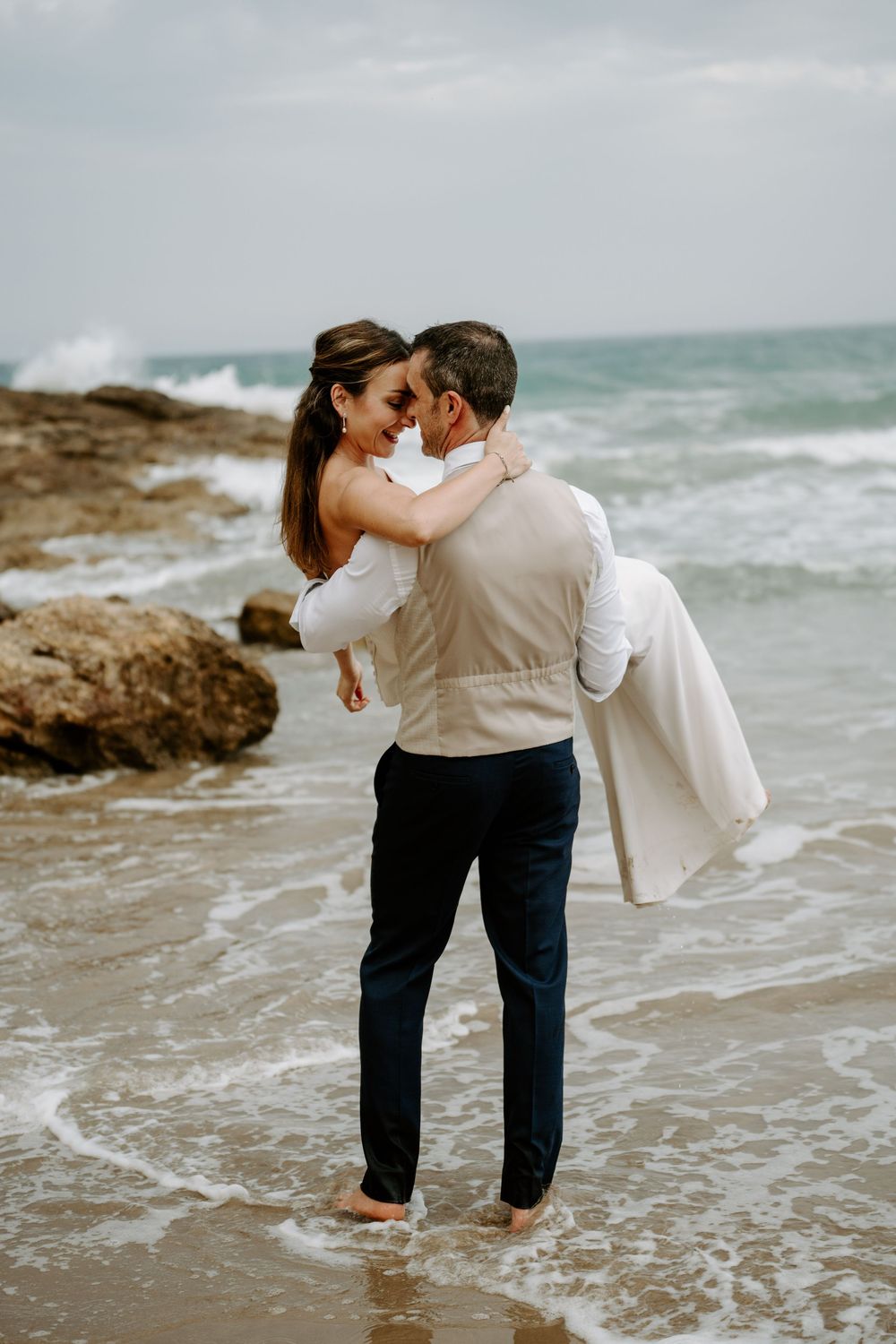Groom in a vest and navy pants carries his smiling bride through shallow ocean waves on a rocky beach.