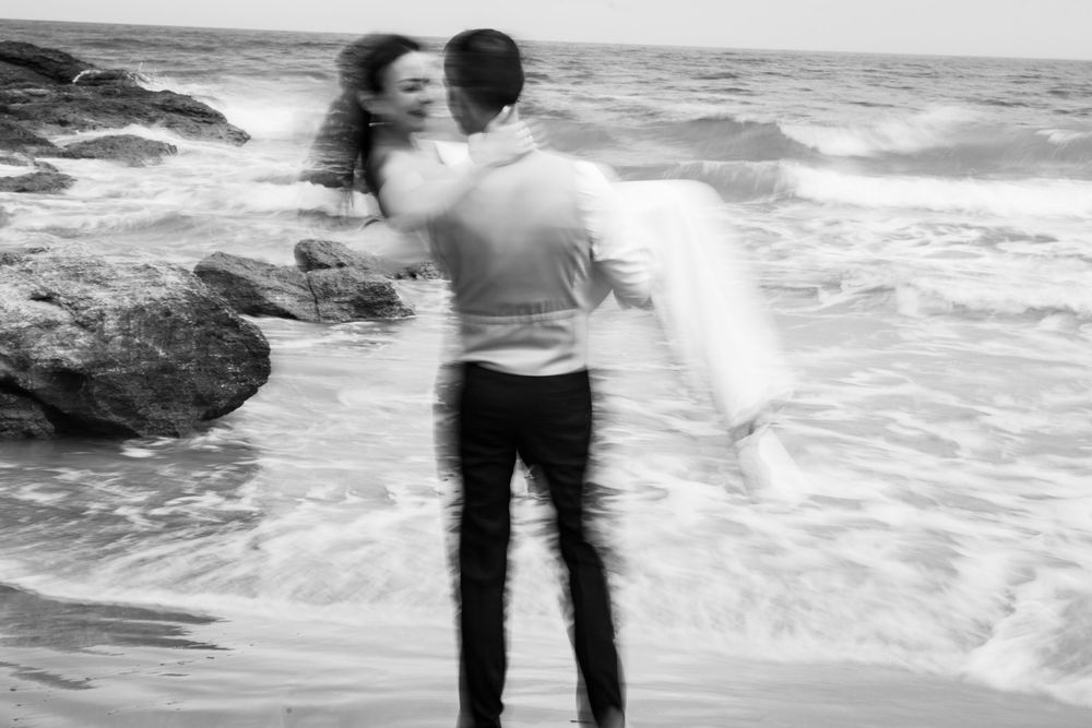 Black and white photo of a man in a dress shirt carrying a woman in his arms on a rocky beach as ocean waves roll in.