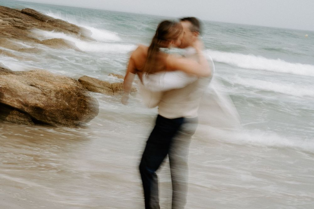 Couple embracing and spinning on a sandy beach with soft motion blur, ocean waves and rocks in the background.