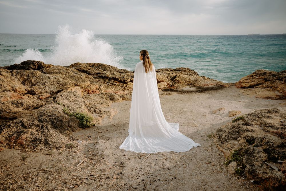 Bride in a flowing white gown and long veil stands on rocky coastline facing the turquoise sea as a wave crashes in the distance.