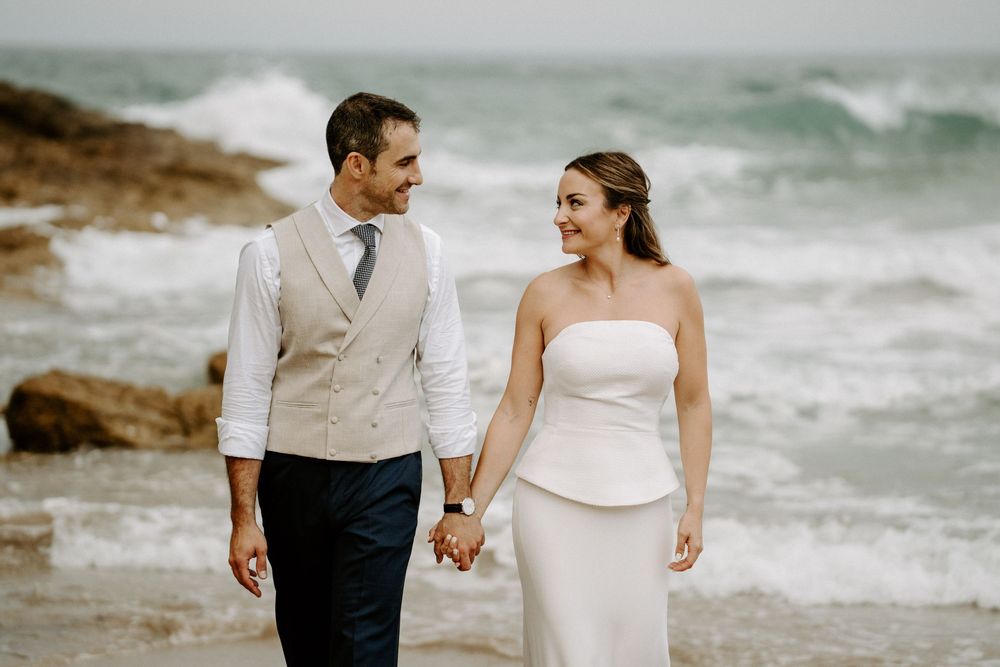 Smiling bride and groom hold hands and gaze at each other on a beach with ocean waves crashing behind them.