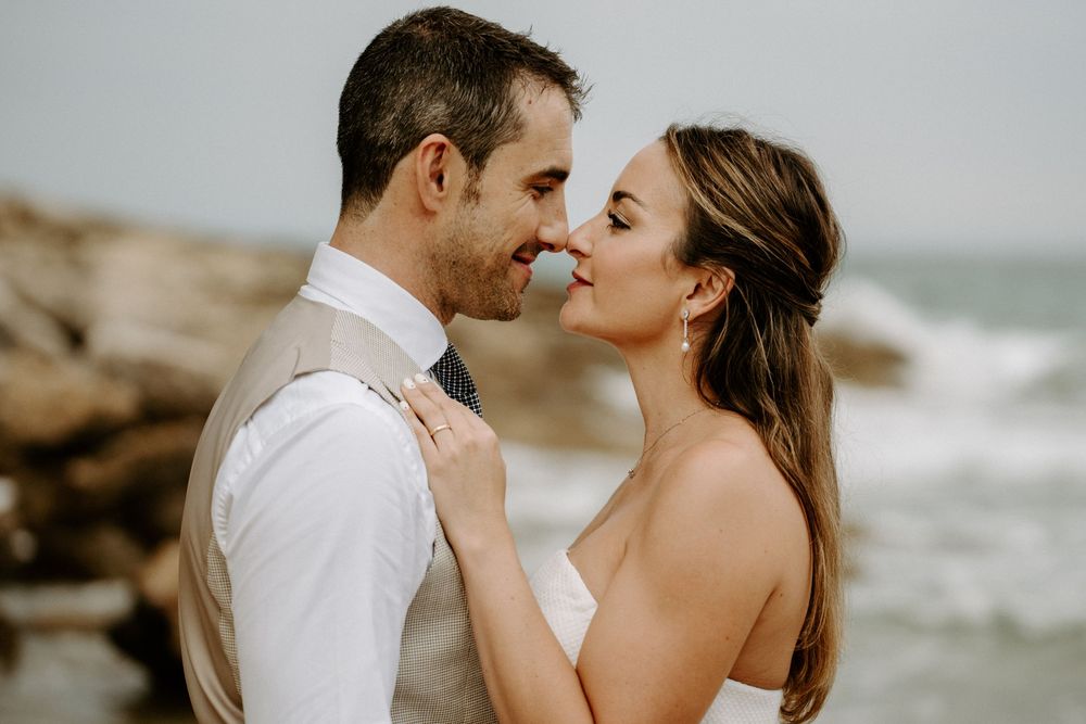 Bride and groom share an intimate moment nose to nose on a rocky beach with ocean waves crashing in the background.