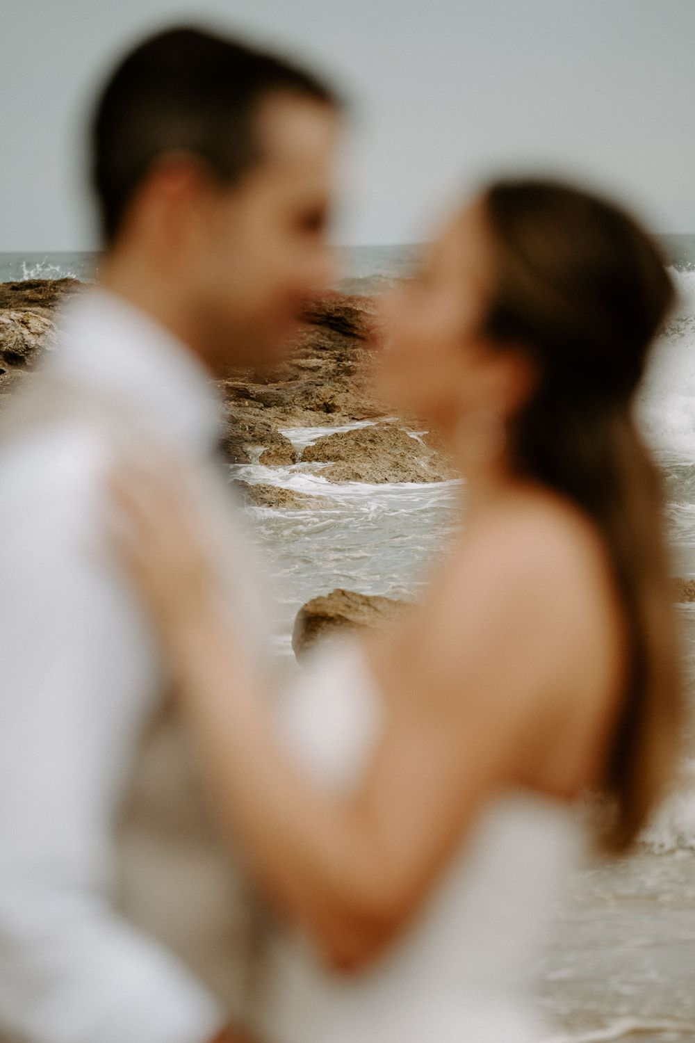 Blurred close-up of a couple in white embracing face to face on a rocky beach with ocean waves in the soft-focus background.