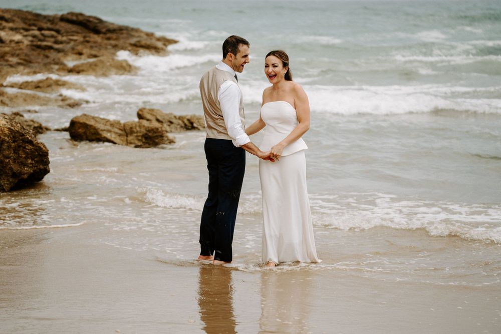 Laughing newlywed couple stand barefoot in shallow seaside waves, holding hands during a joyful beach wedding photo session.