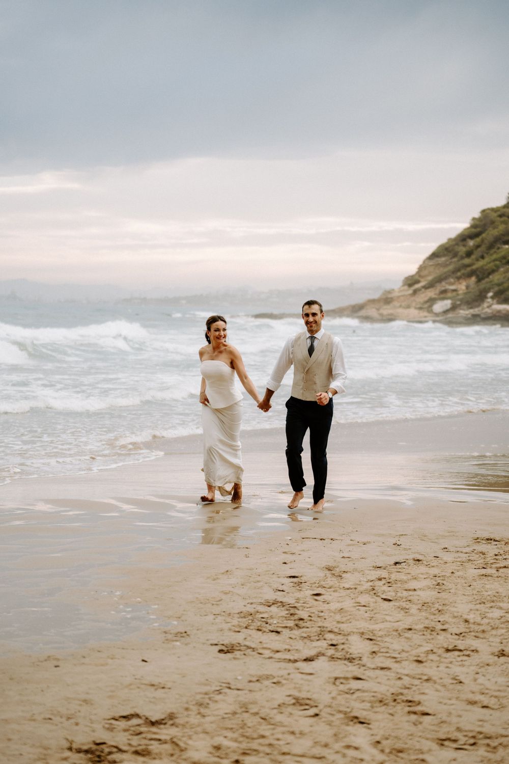 Barefoot bride and groom hold hands and walk along a wet sandy beach with waves and a green headland behind them.