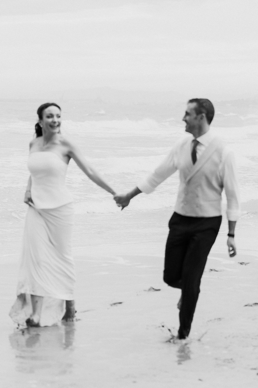 Black and white photo of a smiling bride and groom holding hands as they run through shallow ocean waves on a beach.