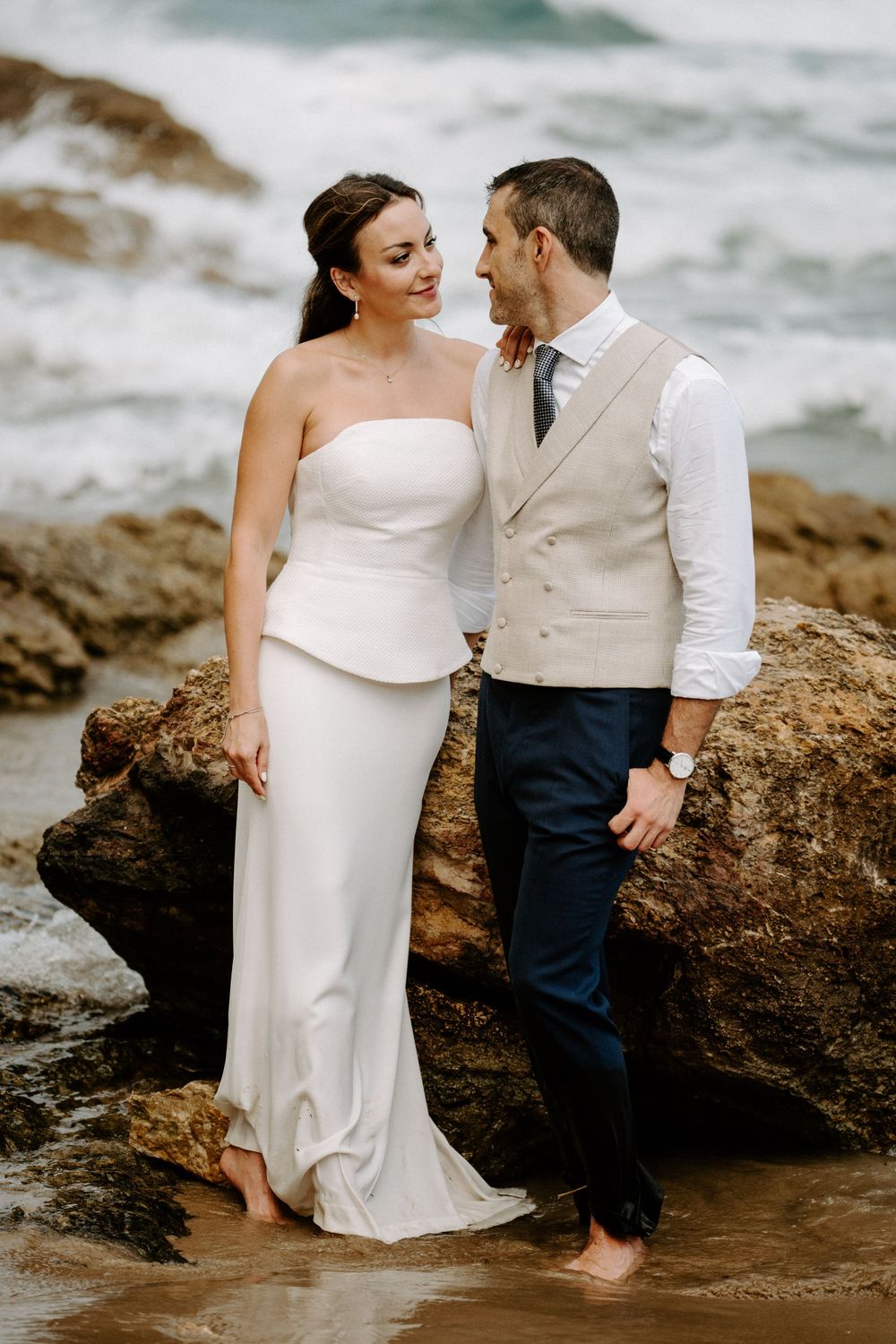 Bride in a strapless white gown and groom in a vest and navy trousers embrace barefoot on rocks by the sea.