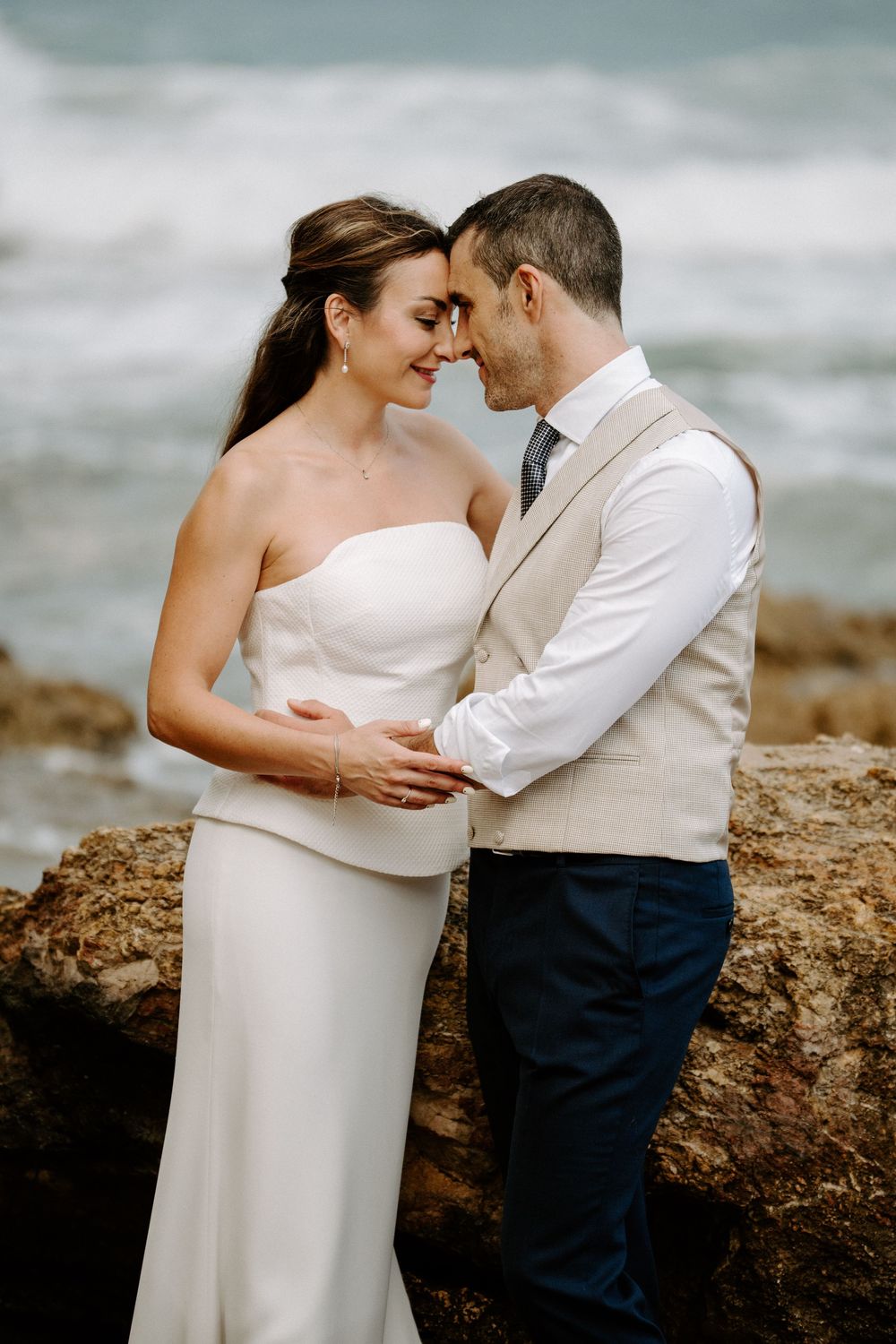 Bride and groom touch foreheads in a tender embrace on a rocky shoreline with ocean waves in the background.