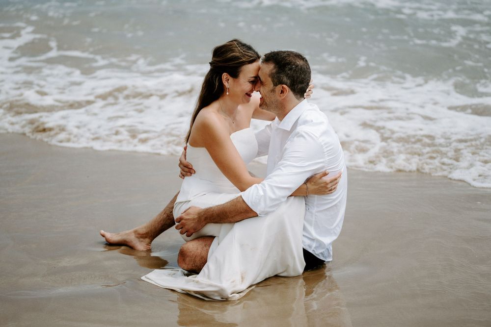 Barefoot couple in white wedding attire embrace and share an intimate moment while sitting in the shallow surf on the beach.