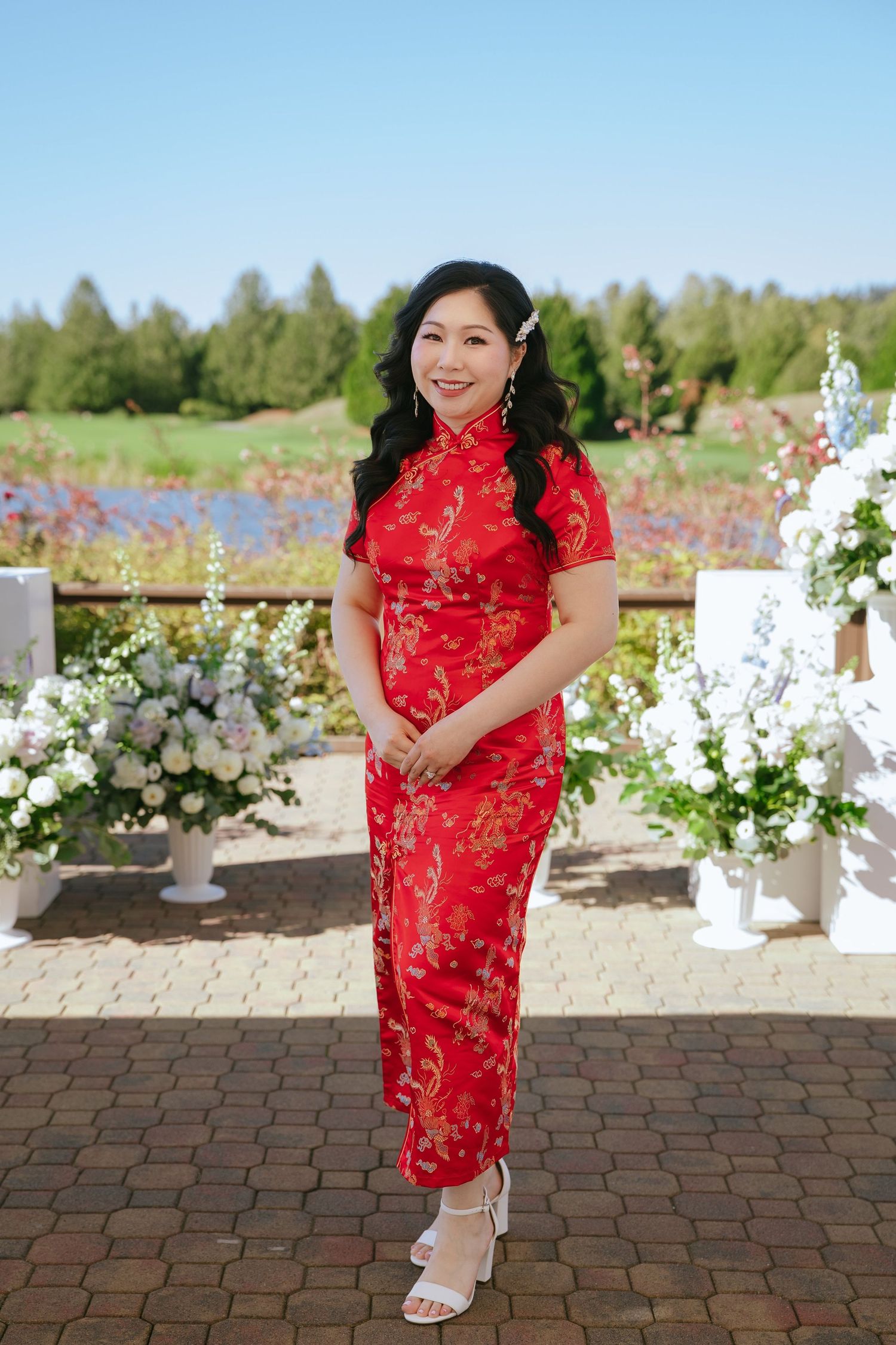 Woman in a red floral qipao and white heels stands on a brick patio beside white flower arrangements at an outdoor event near a pond.