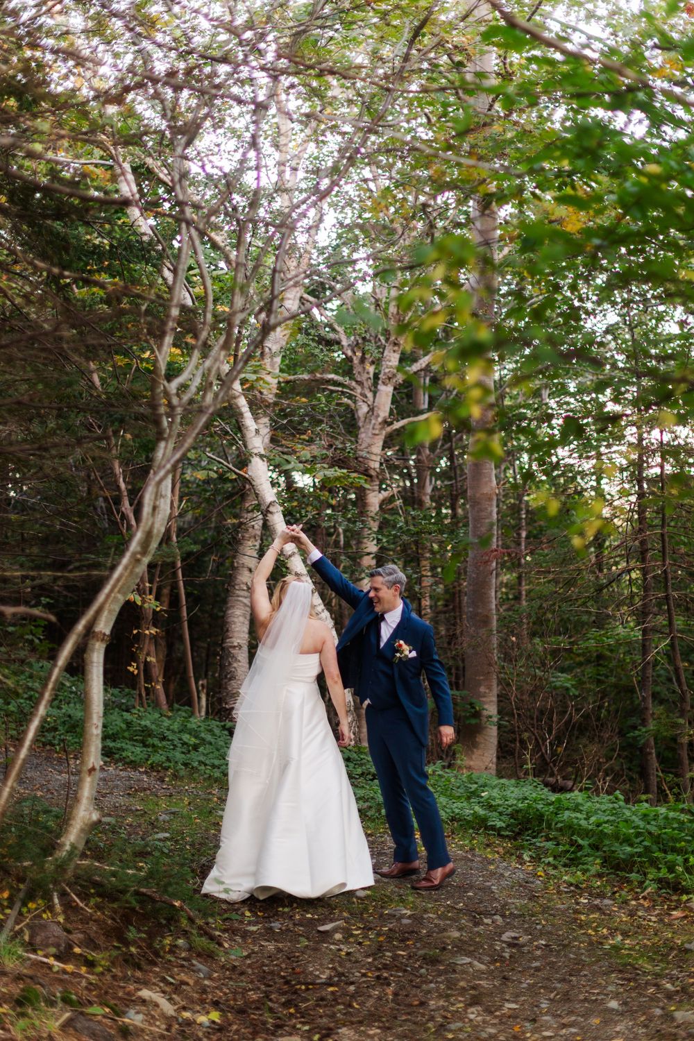 Groom in a navy suit twirls his bride in a white gown along a wooded path surrounded by autumn trees.