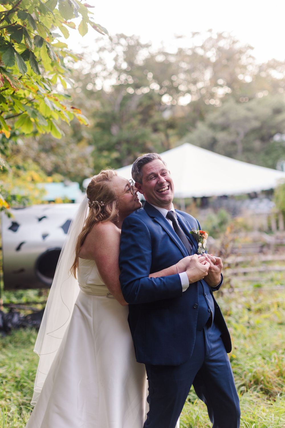 Smiling bride in a white gown and veil embraces her laughing groom in a navy suit during an outdoor garden wedding with a white tent behind.