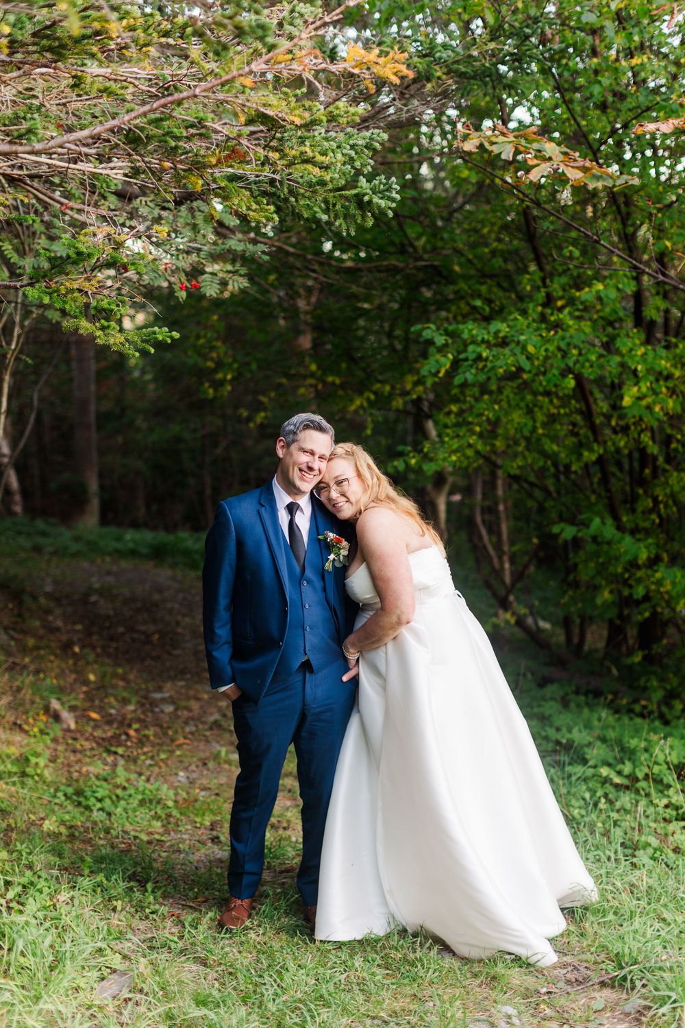 Smiling bride in a white gown leans against groom in a blue suit and tie as they pose for wedding photos among green trees outdoors.