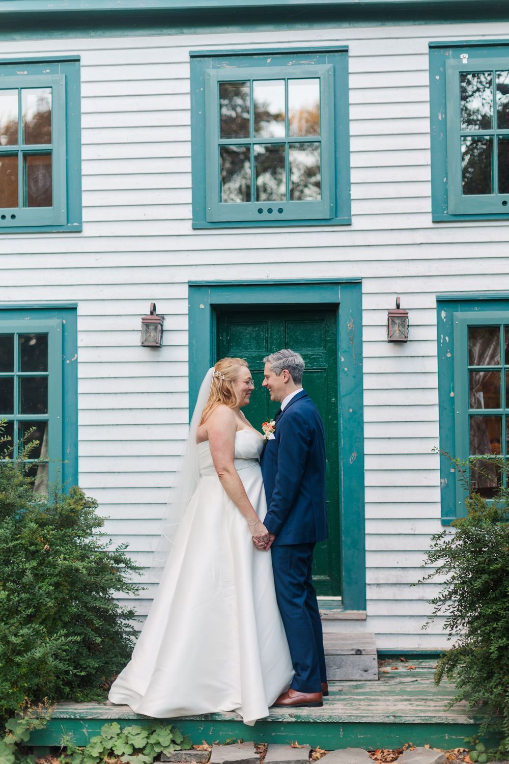 Bride in a white gown and veil holds hands with her groom in a navy suit in front of a white house with a green door.