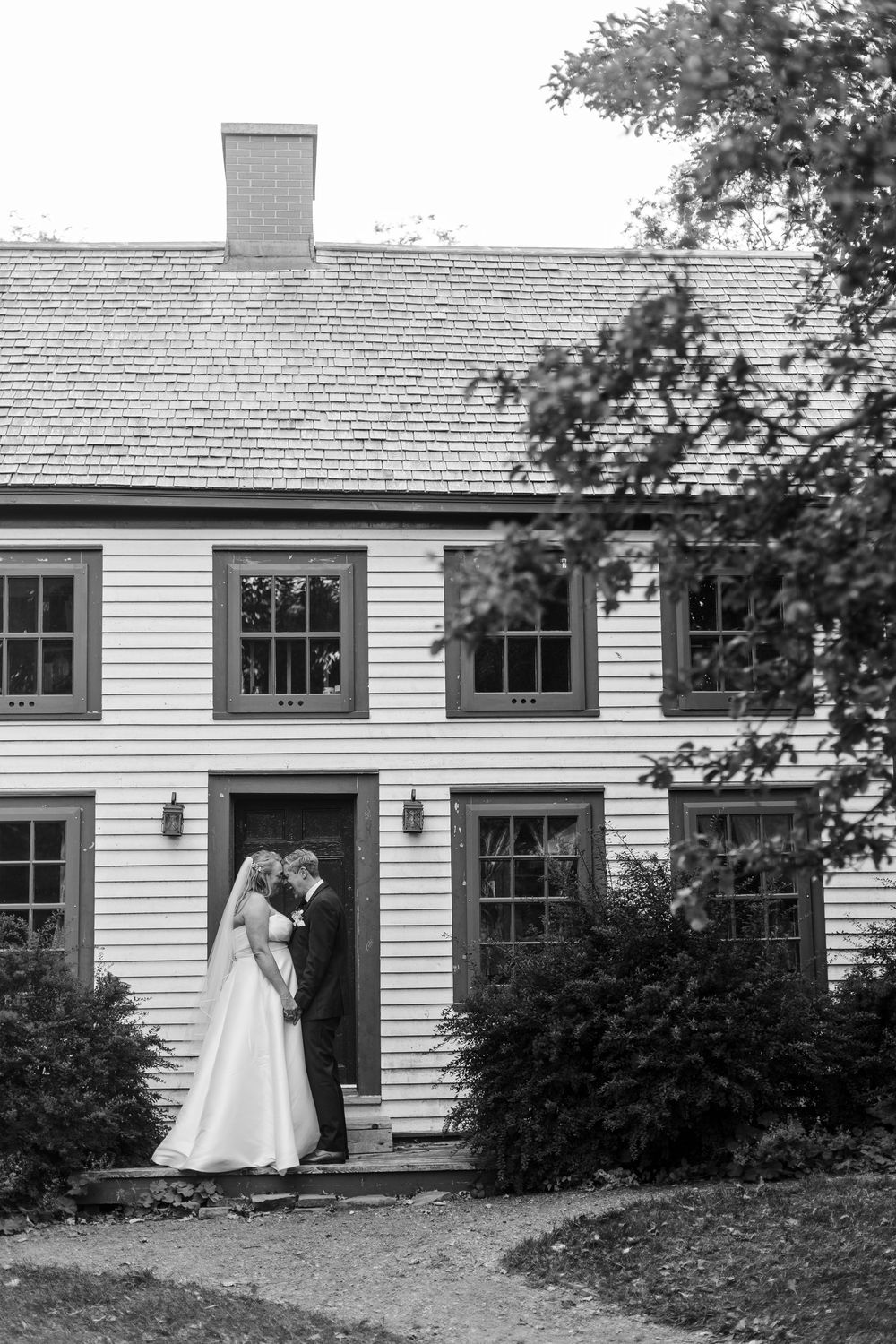 Black and white photo of a bride and groom embracing at the doorway of a historic colonial-style clapboard home with a shingled roof.