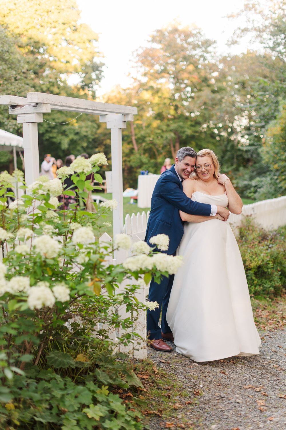 Smiling bride in a strapless white gown embraces her groom in a navy suit beside white hydrangeas and a garden arbor.