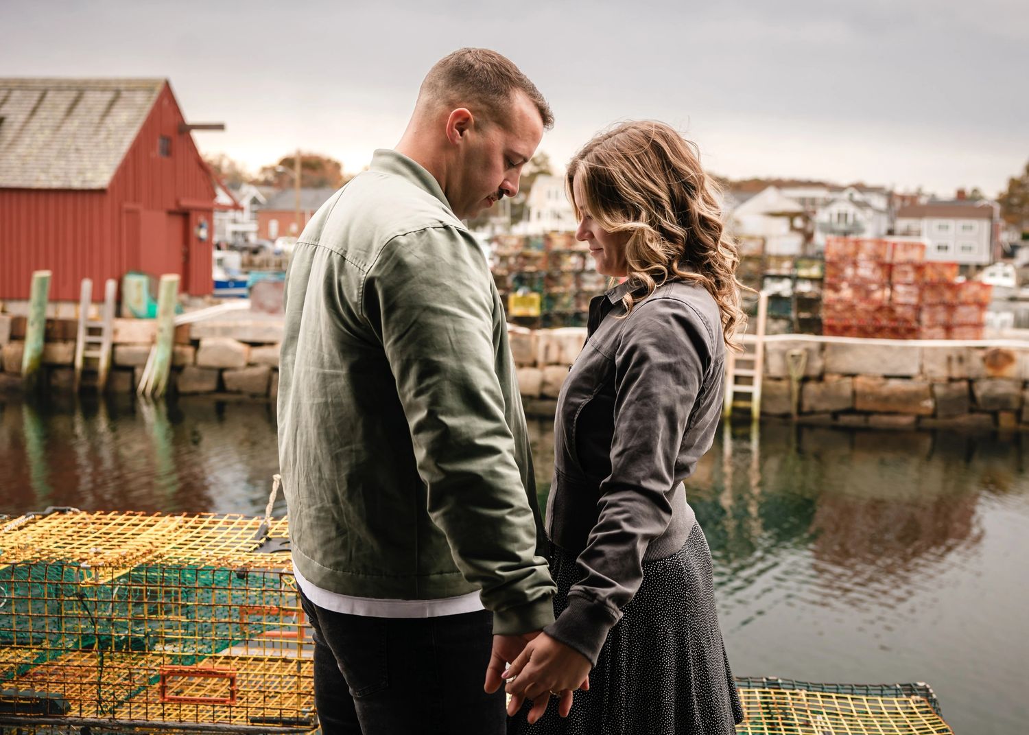 Couple holding hands while standing on a dock overlooking the water, gazing toward the horizon together.  Engagement at Rockport, MA
