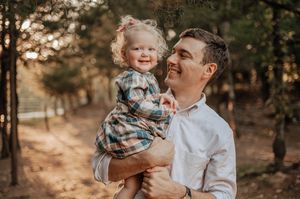 Smiling man in a white button-up shirt holding a laughing toddler girl in a plaid dress outdoors among sunlit trees.