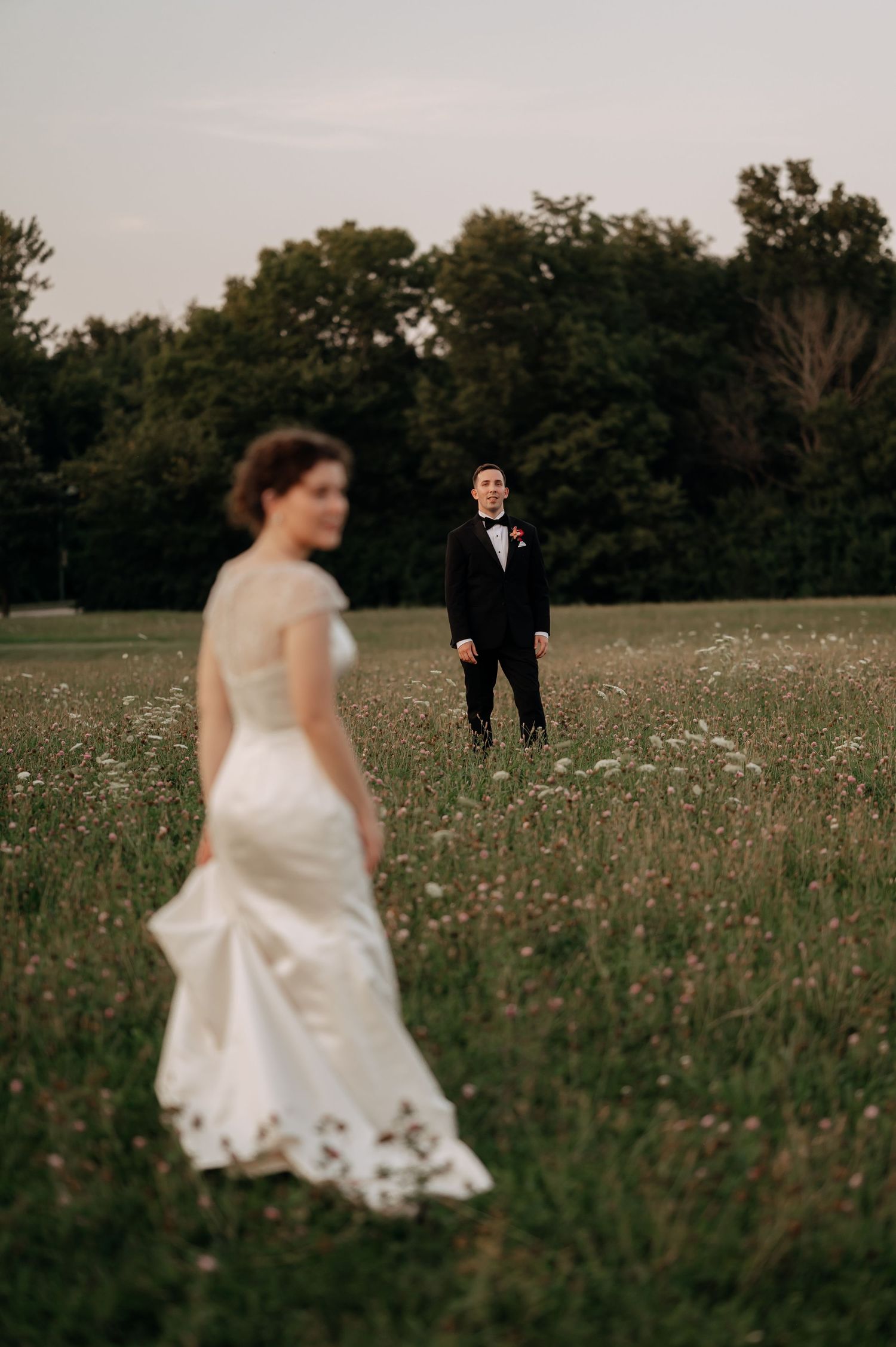 Bride in a white lace wedding gown walks through a wildflower meadow toward the groom in a black tuxedo standing under trees at sunset.