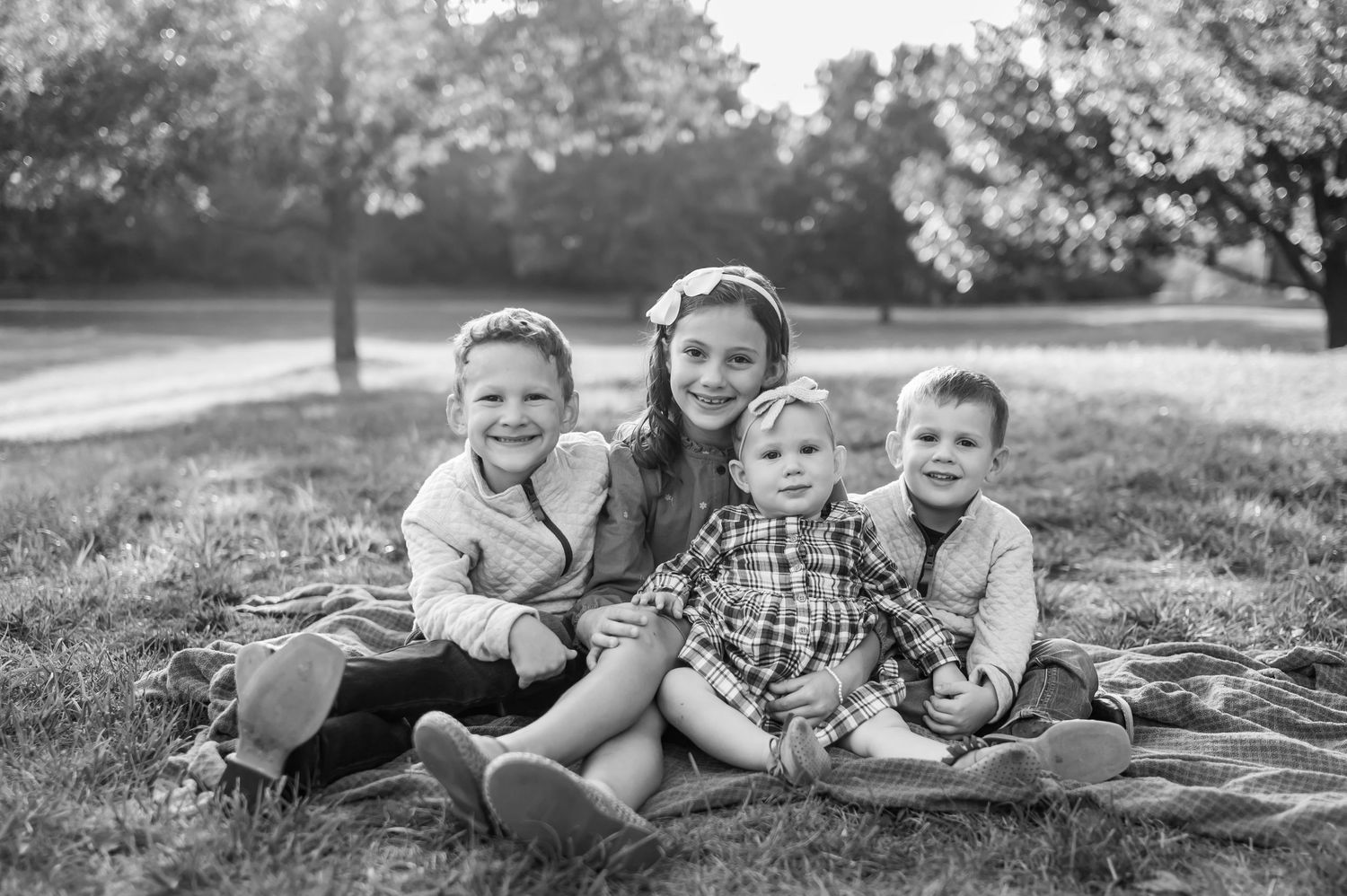 Four smiling children sit close together on a blanket in a grassy park beneath blooming trees in a black and white family portrait.