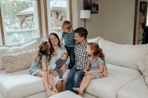 Smiling family of six with a baby, a toddler, and two girls gathered together on a cozy cream sectional sofa in a bright living room.