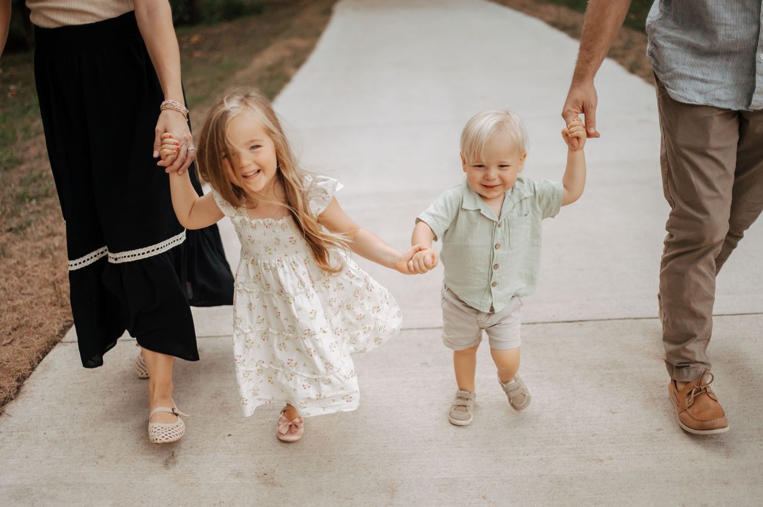 Two smiling toddlers hold hands while walking down a sunny concrete path between two adults in casual summer outfits.