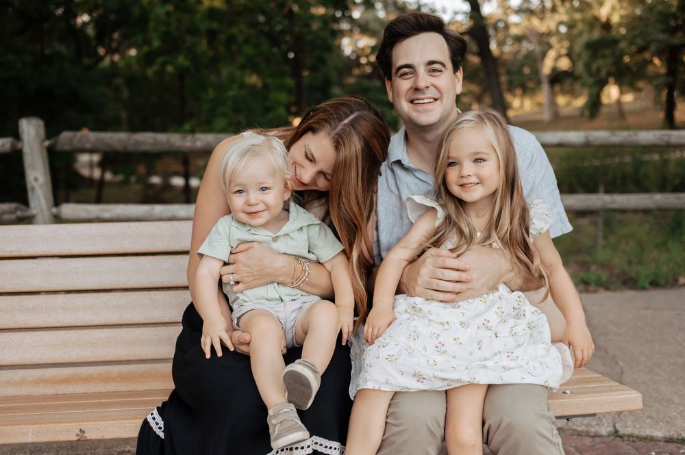 A smiling family of four sits together on a wooden park bench, with two young children on their parents' laps, surrounded by green trees.
