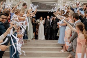 Bride in white gown and groom in dark suit smile on church steps as wedding guests wave white ribbon wands in celebration.