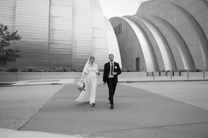 Black and white photo of a bride and groom walking hand in hand outside a modern curved metallic building on their wedding day.