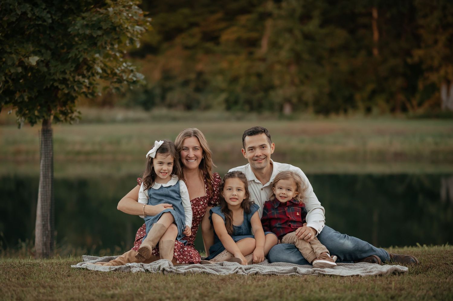 Smiling family of five with three young girls sitting together on a blanket on a grassy lawn beside a calm lake at sunset.