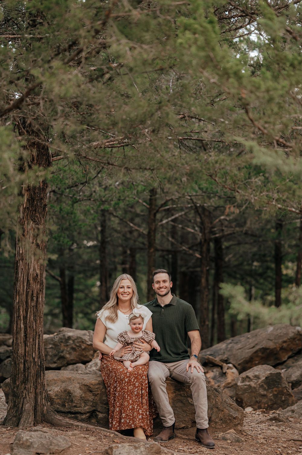 Smiling couple sits on a boulder holding their baby in a sunlit pine forest, a warm outdoor family portrait.