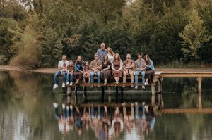 Large family of twelve posing together on a wooden dock over calm water, surrounded by green trees and reflected in the lake.