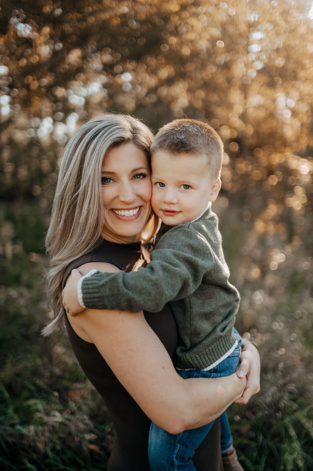 Smiling woman with long blonde hair holds a young boy in a green sweater and jeans outdoors in warm golden sunset light.