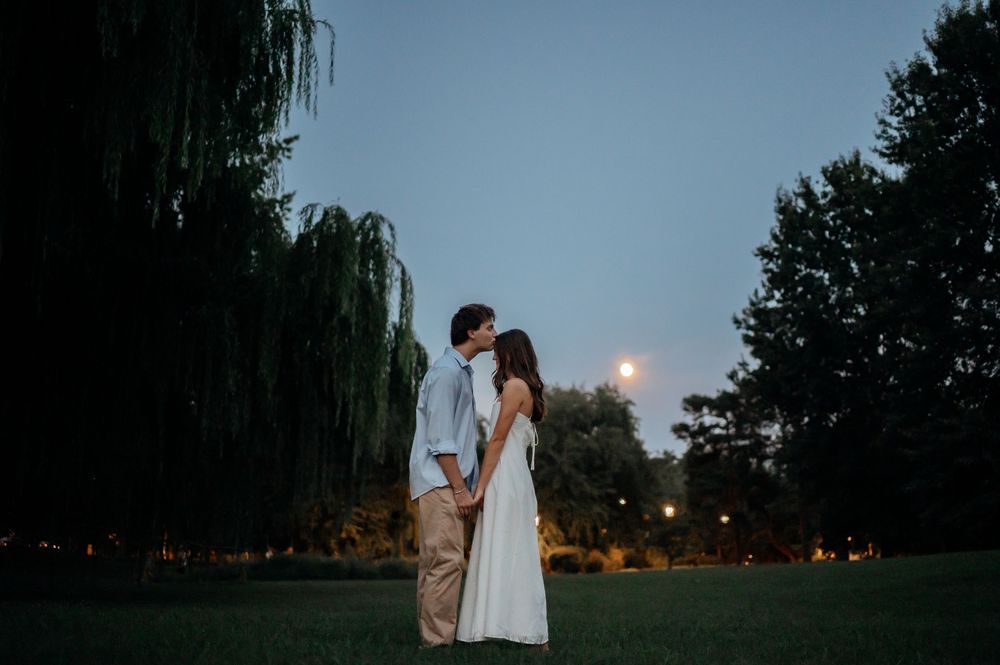 Couple in light summer clothes share a kiss on a grassy lawn at dusk, framed by a weeping willow and a glowing moon.