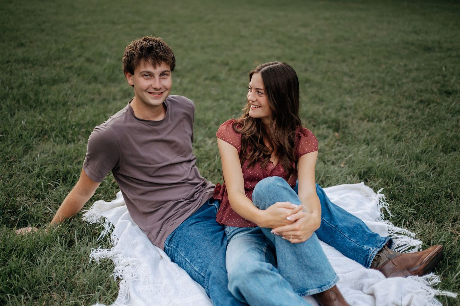 A smiling young man and woman sit close together on a white blanket in a green grassy field, both wearing jeans and casual tops.