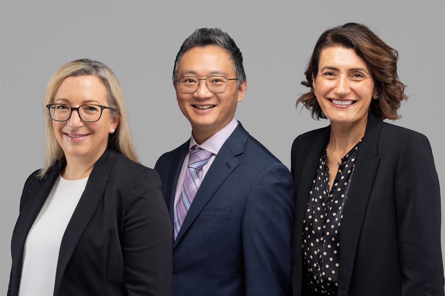 Three professional business people in formal attire posing for a corporate headshot on grey background.