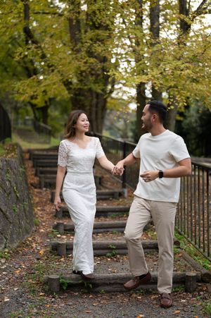 Smiling couple holding hands and walking down wooden park steps beneath autumn trees, she in a white lace dress, he in a tee and khakis.