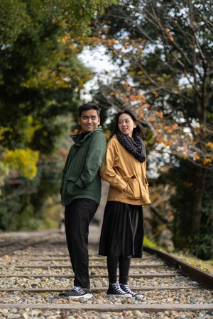 Smiling couple standing back to back on railway tracks, surrounded by colorful autumn trees.
